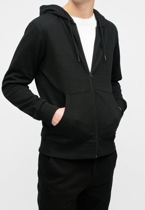 Mand iført en sort hættetrøje med lynlås og hænder i forlommerne, over en hvid T-shirt, stående mod en ensfarvet lys baggrund.