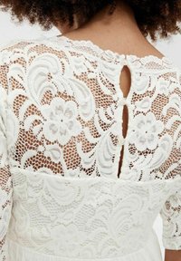 Robe en dentelle blanche ornée de motifs floraux, fermée par des boutons au dos, et dotée d'une jupe en tissu lisse avec des manches en dentelle transparente.