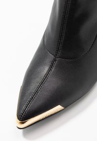 Czarny skórzany but z ostrym noskiem, z złotymi akcentami na obcasie. Gładka tekstura z widocznymi detalami szwów.