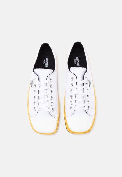 Ζευγάρι λευκά δερμάτινα sneakers Moschino με κίτρινες σόλες, μαύρο εσωτερικό επένδυμα και εμφανές όνομα επωνυμίας στις εσώσολες και στα πλάγια.