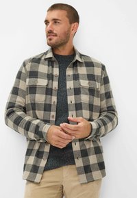 Bisonternet skjorte i beige og sort flannel, med to frontlommer og knaplukning. Båret over en mørk struktureret sweater.