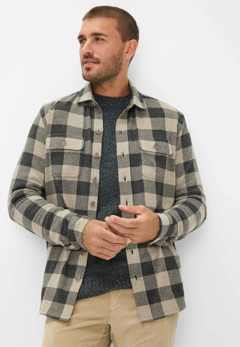 Bisonternet skjorte i beige og sort flannel, med to frontlommer og knaplukning. Båret over en mørk struktureret sweater.