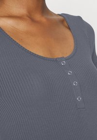 Primer plano de una persona que lleva una blusa de canalé gris oscuro con escote amplio y cuatro pequeños botones de presión plateados en la parte frontal.