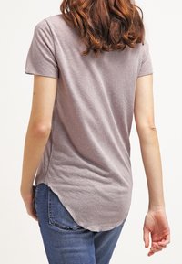 Kortärmad, ljusgrå T-shirt med rundad nederkant och fransade kanter, kombinerad med blå jeans. Tyget ser mjukt och avslappnat ut.