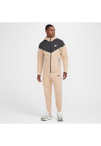 Completo sportivo beige e grigio con felpa con zip e pantaloni a sigaretta; presenta il logo Nike, colori a contrasto e tasche laterali.