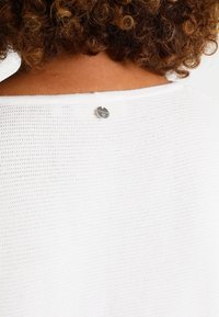 Suéter blanco de punto con un patrón texturizado, que presenta un pequeño logo plateado en la parte posterior del escote.