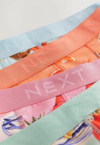 Vier farbenfrohe Gummibänder mit dem eingeprägten Text "NEXT": hellblau, korallenrot, rosa und mintgrün, die über einem Blumenmuster liegen.