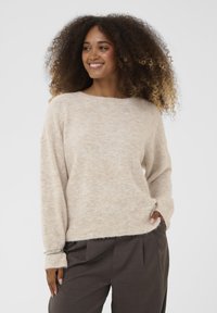 Bež, teksturirani pulover s opuštenim krojem, širokim okruglim izrezom i dugim rukavima. Uparen s tamnim, nabranim hlačama. Vidljiva suptilna dlačica.