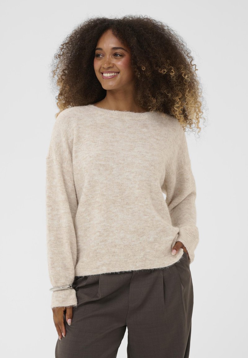 Bež, teksturirani pulover s opuštenim krojem, širokim okruglim izrezom i dugim rukavima. Uparen s tamnim, nabranim hlačama. Vidljiva suptilna dlačica.