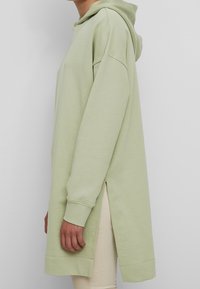 Felpa verde chiaro, realizzata in tessuto morbido, presenta una forma lunga, fessure laterali e polsini a costine. Ha un cappuccio con coulisse per una vestibilità regolabile.