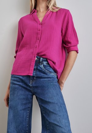Vrouw draagt een felroze, gestructureerd overhemd met opgerolde mouwen en een blauwe spijkerbroek, haar hand in de zak gestoken tegen een effen achtergrond.