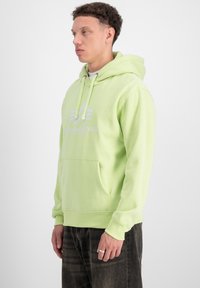 Felpa verde chiaro con cappuccio e coulisse, tasca a marsupio e logo frontale. Il tessuto sembra morbido con una superficie liscia.