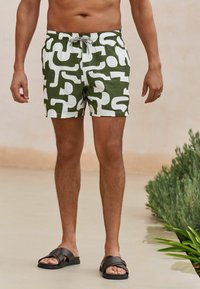 Green and white patterned swim shorts with an elastic waistband and drawstring. Worn with brown slide sandals. Textured fabric.