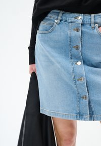 Ljusblå knappframdressad denimkjol som bärs med en svart topp, och en hand som håller en svart läderjacka vid sidan.