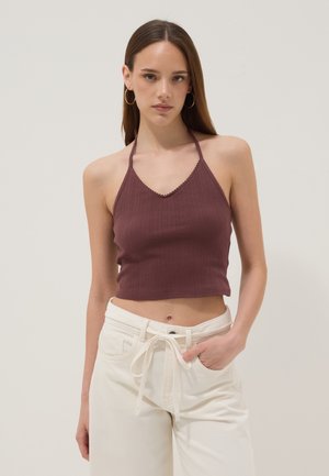 Jonge vrouw draagt een mouwloze bordeauxrode halter crop top en witte hoge taille broek met een gestrikte riem, staand tegen een effen achtergrond.