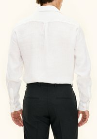 Camicia bianca di lino con maniche lunghe e polsini con bottoni; abbinata a pantaloni grigio scuro su misura con due tasche posteriori e dettagli sartoriali sottili.