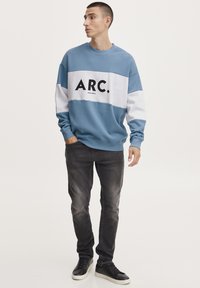 Felpa azzurra chiara con strisce orizzontali bianche e blu scuro, caratterizzata dalla scritta "ARC." in nero. Indossata con jeans scuri e sneaker nere.