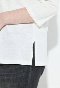Persona che indossa una camicia bianca a maniche corte con una fessura laterale su pantaloni grigio scuro, mostrata dalla parte centrale del torso fino alla parte superiore della coscia.