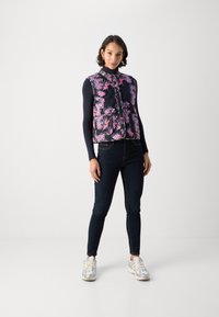 Blumengemusterte Steppweste in Schwarz und Pink, mit hohem Kragen und einer Knopfleiste vorne. Trägt man über einem Langarmshirt und kombiniert mit dunklen Jeans.