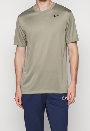 Homme portant un t-shirt Nike vert olive clair à manches courtes et un pantalon Nike bleu marine, debout devant un fond uni.
