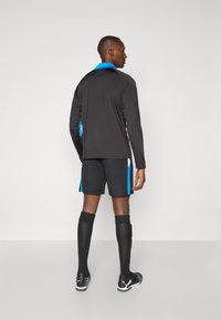 Svart langermet skjorte med blå detaljer, kombinert med svarte shorts med blå sidestriper. Svart knelange sokker og svarte sportssko.