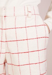 Pantalons à carreaux blancs avec un tissu texturé présentant des lignes rouges, une taille ajustée et des poches latérales. Le motif se compose de grilles horizontales et verticales.