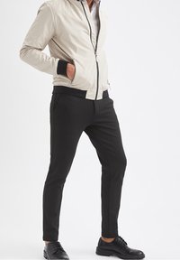 Hellbeige Bomberjacke mit schwarzem Besatz, Reißverschluss vorne und zwei seitlichen Taschen, kombiniert mit schwarzen, zulaufenden Hosen und schwarzen Schnürschuhen.