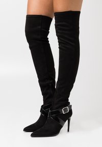 Botas negras de ante hasta la rodilla con punta afilada, hebilla de charol y tacón de aguja. Detalle de cremallera en el costado.