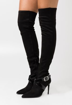 Botas negras de ante hasta la rodilla con punta afilada, hebilla de charol y tacón de aguja. Detalle de cremallera en el costado.