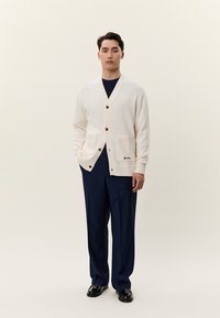 Man står mot en enfärgad bakgrund iklädd en off-white cardigan, marinblå skjorta, marinblå byxor och svarta blanka skor, med händerna avslappnade vid sidorna.