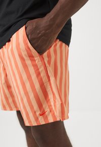 Personne à la peau foncée portant un short à rayures verticales orange et orange clair avec un petit logo Nike rouge, main dans la poche, chemise noire visible.