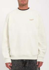Sweatshirt en polaire blanche avec un col rond, des poignets côtelés et une petite étiquette beige sur la poitrine. Texture douce, coupe décontractée.