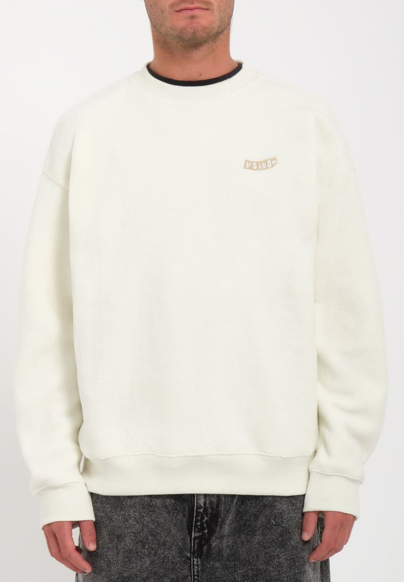 Sweatshirt en polaire blanche avec un col rond, des poignets côtelés et une petite étiquette beige sur la poitrine. Texture douce, coupe décontractée.