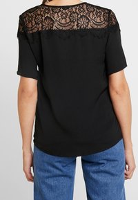 Blusa nera con una sovrapposizione di pizzo sul retro caratterizzata da motivi floreali. La blusa ha maniche corte e un orlo stondato.