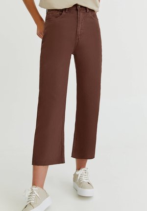 Jean brun coupé droit raccourci, porté avec des baskets beige et un haut beige clair rentré, debout sur un fond clair.