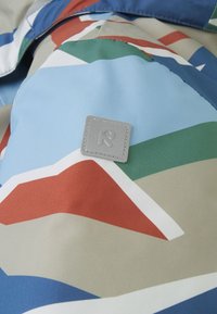 Vodootporna tkanina s geometrijskim uzorcima u plavoj, crvenoj, zelenoj i bež boji. Sivi etiketa s utisnutim detaljem u obliku slova "R".