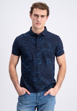 Jonge man met krullend haar die een donkerblauw poloshirt met bloemmotief en lichtblauwe spijkerbroek draagt, handen in de zakken, neutrale gezichtsuitdrukking.