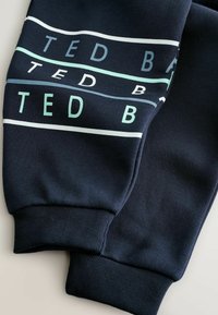 Marineblaue Jogginghosen mit gerippten Bündchen, versehen mit weißen und hellblauen horizontalen Streifen sowie dem Textmuster "TED B" entlang der Beine.