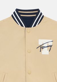 Tan bomber jacket featuring a ribbed collar with navy and white stripes, snap buttons, and a blue embroidered logo on the chest.