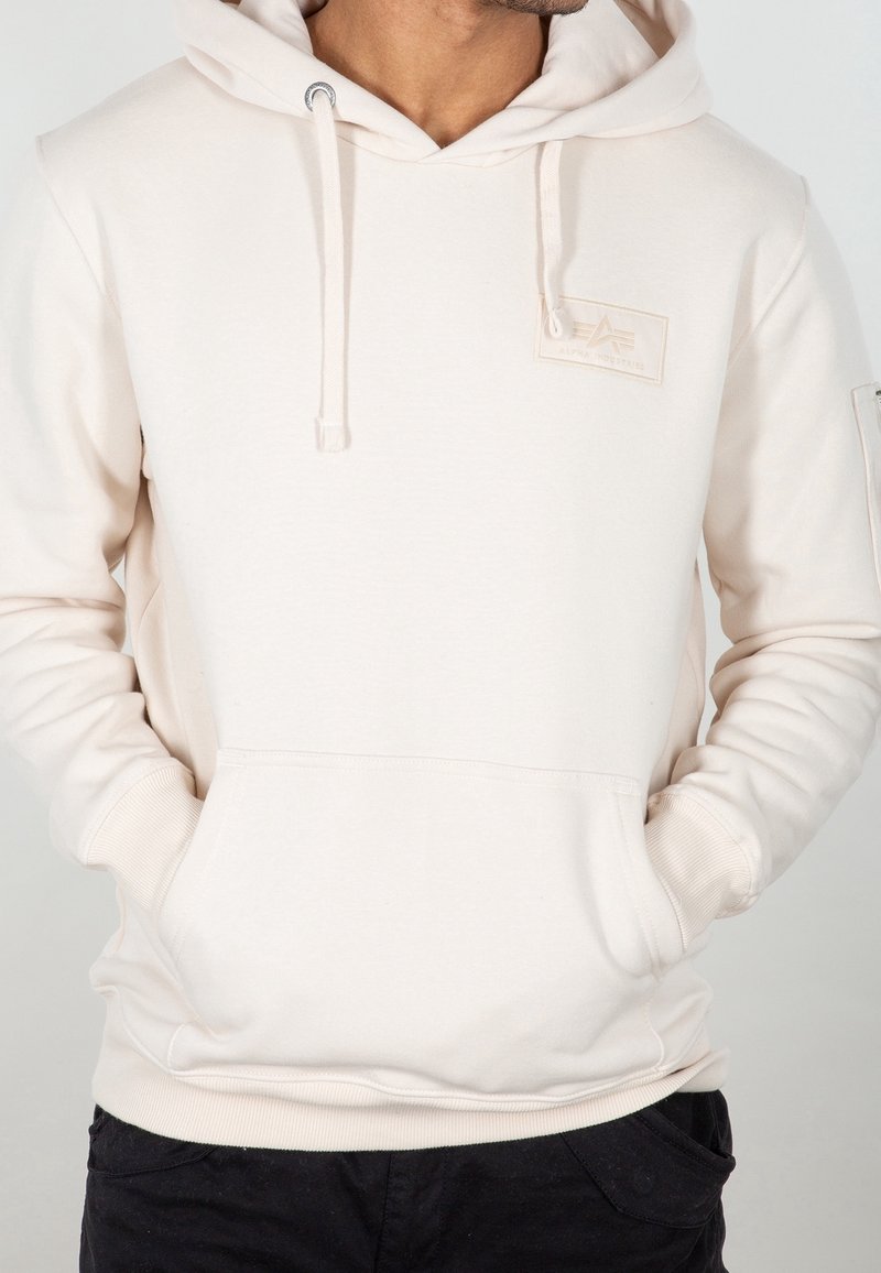 Homme portant un sweat à capuche beige clair avec poche frontale et écusson logo sur le côté gauche de la poitrine, mains dans les poches, cadré sous le menton.