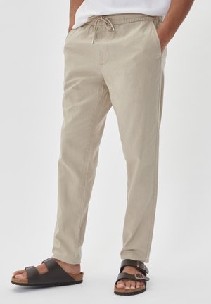 Pantalons légers beige avec taille élastique à cordon, poches latérales et coupe fuselée, associés à des sandales marron à deux brides.