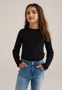 Jeune fille portant un haut noir à manches longues et un jean bleu clair, les mains dans les poches, se tenant devant un fond beige uni.