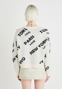 Kremgenser med svart tekst som har bynavn og fraser. Myk, strikket tekstur, ribbet krage og nederkant, crop design.