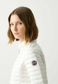 Witte gewatteerde jas met een hoge kraag, voorzien van horizontale stiksels en een ronde Colmar-logo patch op de linker mouw. Gladde textuur.