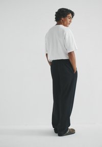 Weißes Kurzarm-T-Shirt kombiniert mit locker sitzenden schwarzen Hosen und braunen Slippern. Model steht mit Händen in den Taschen, von hinten betrachtet.