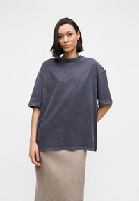 Επιστημονικό γκρι T-shirt σε υπερμεγέθη γραμμή, με στρογγυλή λαιμόκοψη, κοντά μανίκια και μαλακό, υφασμάτινο ύφασμα με υφή. Φοριέται πάνω από μια ανοιχτό γκρι φούστα.