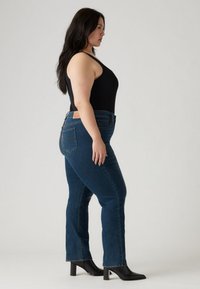 Tamsiai mėlynos spalvos džinsai plačiais klostėtais kojomis ir aukštu liemeniu, turintys kišenes gale bei odinį žymeklį juosmens juostoje. Derinami su juodu be rankovių viršutiniu drabužiu ir juodais aukštakulniais.