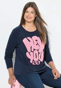 Sudadera azul marino de manga larga con un gráfico rosa de felpa que dice "HEY YOU". Tejido suave, corte relajado, combinada con jeans oscuros.