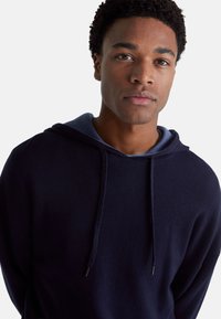 Marineblauwe hoodie met een trekkoord, voorzien van een zachte textuur en gelaagd ontwerp met een lichterblauwe binnenkant. Geen zichtbare logo's of patronen.