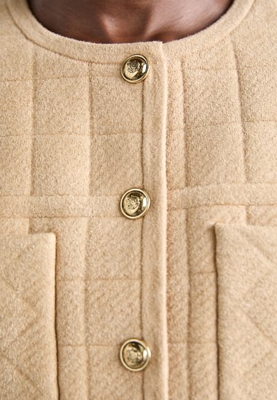 Μπεζ υφασμένη ζακέτα με τρία χρυσά διακοσμητικά κουμπιά και δύο μπροστινές τσέπες.